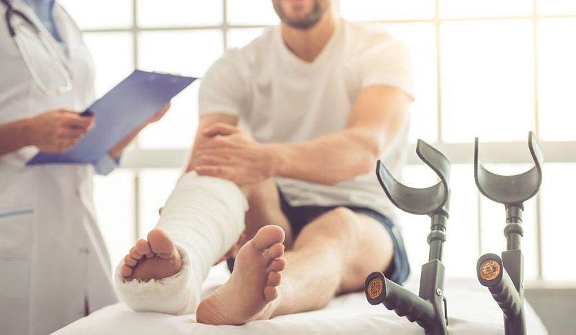 Cropped image of beautiful female medical doctor listening to handsome patient with broken leg and making notes while working in her office