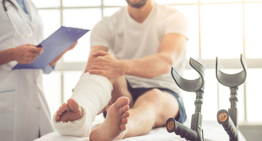 Cropped image of beautiful female medical doctor listening to handsome patient with broken leg and making notes while working in her office